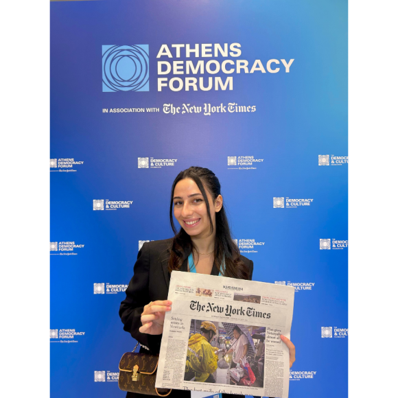 Laila Khalifa standing in front of a Athens Democracy Forum banner holding a copy of the newspaper the New York Times