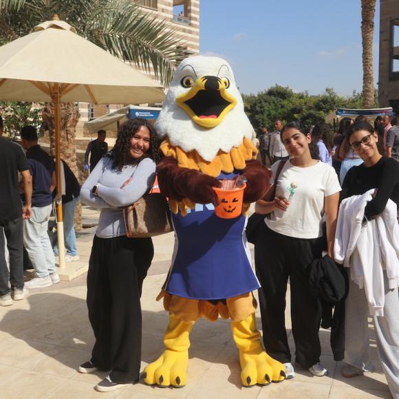 The outdoor on campus is packed with people moving across different booths. At the forefront, poses 3 girls with AUC's eagle mascot. The mascot, wearing a navy jersey reading AUC, stands with his arms out holding an orange pumpkin candy basket. Two girls are on his right dressed in sweatpants and tilting their heads towards the mascot. The girl on his left poses with her arms crossed, with her back leaning on the mascot and smiling.