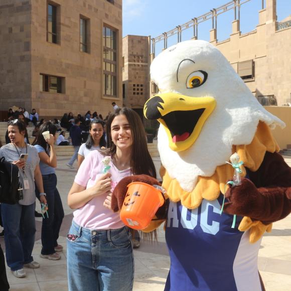 The photo is taken in the outdoors of campus. the background is flooded with students walking around and hanging out. At the face of the image is a female student with long, dark hair, wearing a pink shirt and jeans while holding a cake pop. She poses right next to the AUC mascot; an eagle costume is seen with a white and yellow head of the eagle, his yellow beak and eyes, and a happy expression. He wears a blue jersey saying "AUC" holding an orange pumpkin basket and a cake pop.