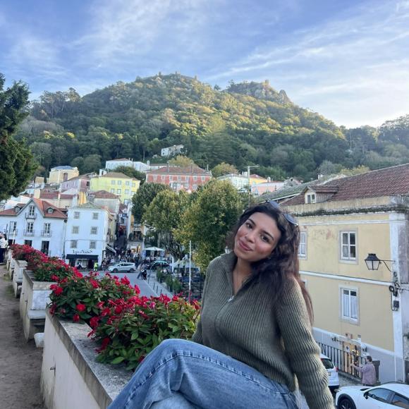 A female is sitting beside flowers and there are houses and a green hill behind her